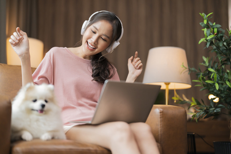young asian cheerful freshness Woman waving while she is conversation during video call and wear headphone device using laptop while her white hair colour pomeranian dog pet is relax laying down sit next to her on sofa armchair at home