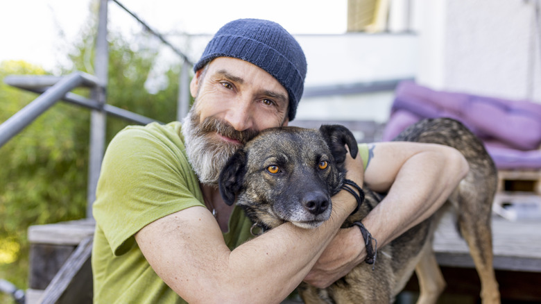 A bearded man sits on a porch and hugs his dog that is standing next to him