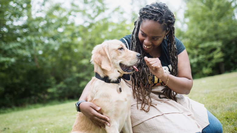 A smiling woman kneels beside her dog, hiding a treat in the closed hand she is holding in front of the dog's face