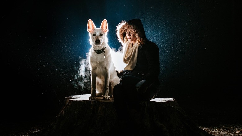 Woman in a hooded jacket sits beside a white shepherd dog against a dark background.