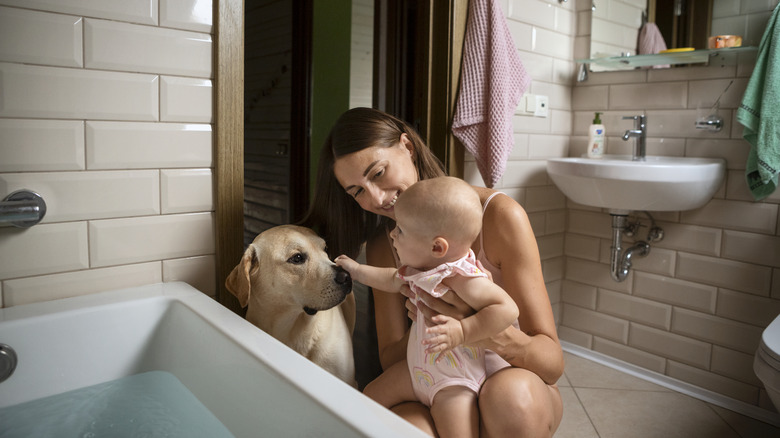 A woman holds a baby as it boops the nose of a Labrador retriever near a bathtub with water in it