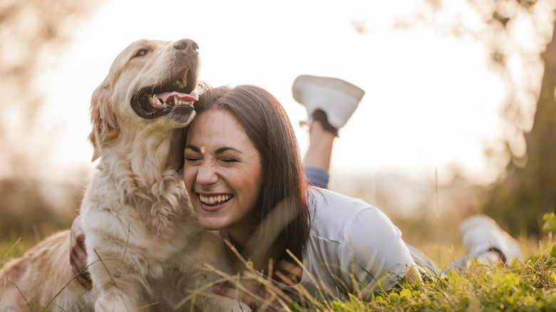 A smiling woman lying in a field and hugging a golden retriever.