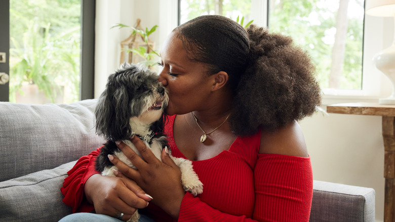 A curly-haired woman in a red shirt hugging and kissing a Havanese dog.