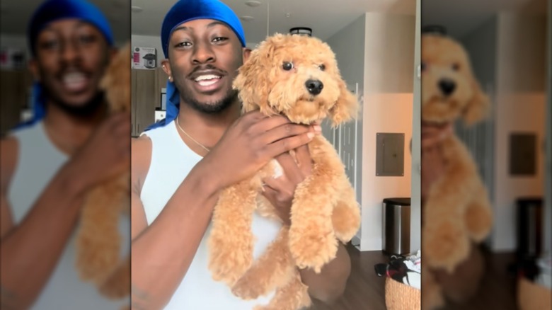 A man in a white tank top and blue durag carrying a mini poodle.