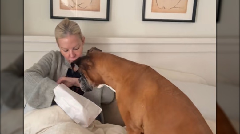 A woman reaching into a white bag while a dog watches.