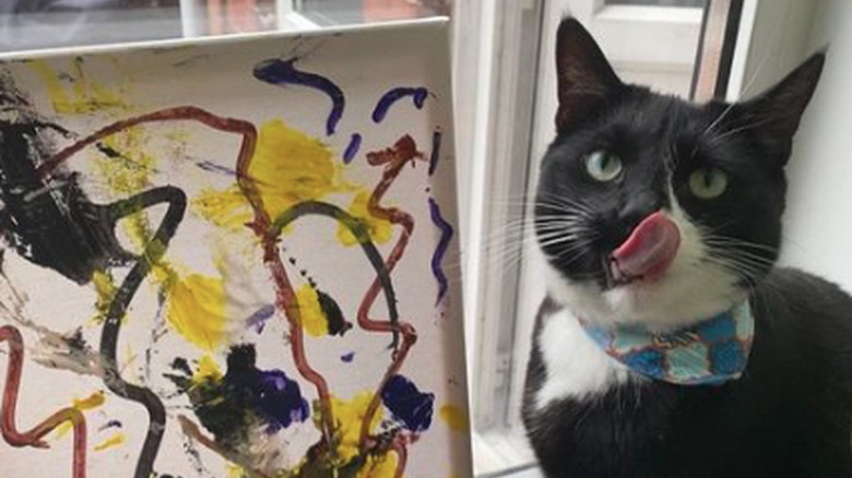 A small painted canvas resting in a windowsill. Next to the painting sits a cat with their tongue out, licking their nose.
