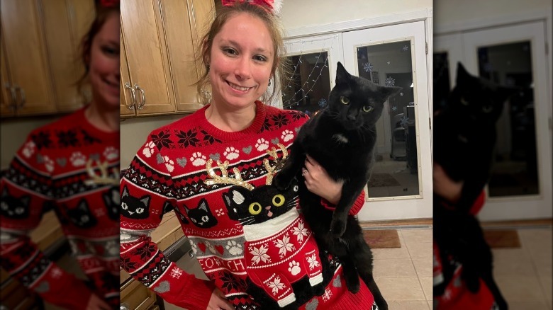 A woman is wearing a Christmas sweater with a black cat on it, while holding her own black cat.