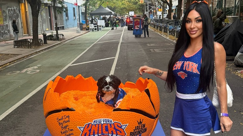 Shih Tzu dressed as Knicks player Jalen Brunson inside a giant bright orange basketball on a parade float beside a white woman in a Knicks dancer costume on New York street.
