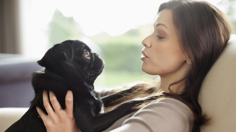 White woman with long brown hair sits while holding a black pug and talking to it.