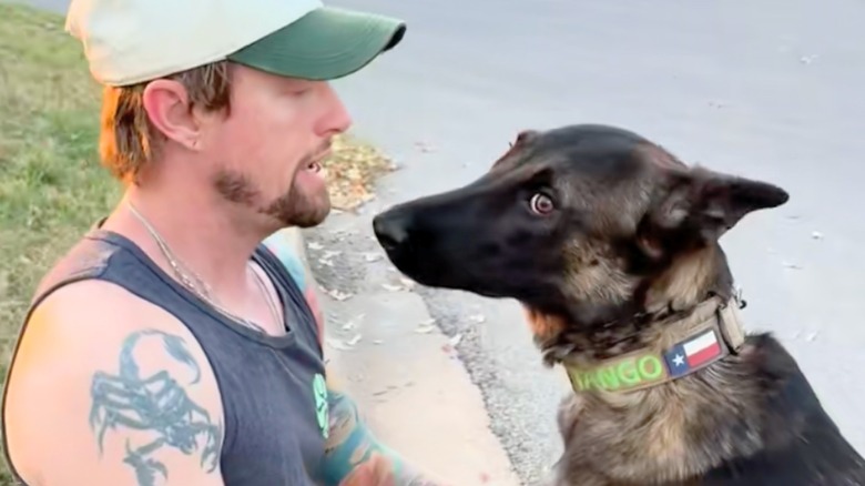 Belgian Malinois looks away as man in grey tank top and tan and white hat speaks.