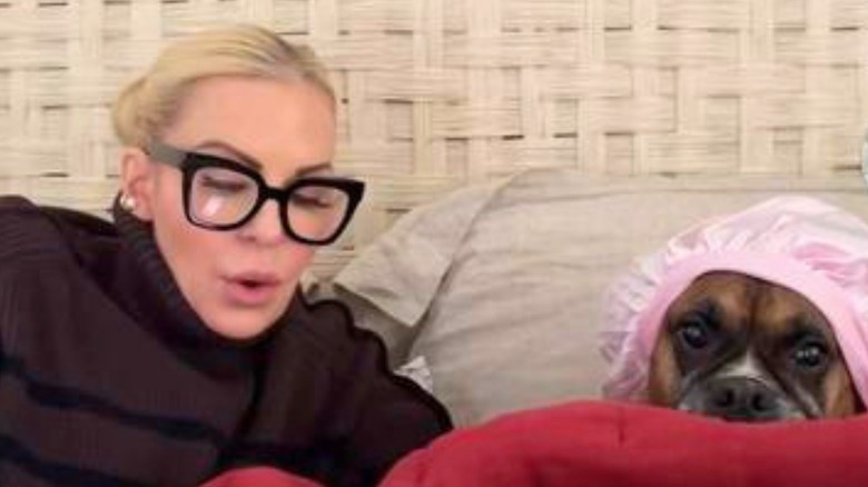 Grandma with glasses and blonde hair in a bun reads to a boxer wearing a pink bonnet while the two lay in bed under red cover.