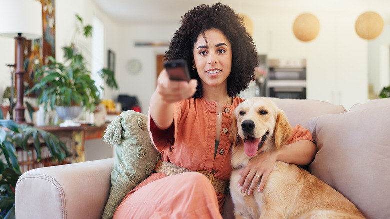Woman uses TV remote and relaxes with dog on tan couch.
