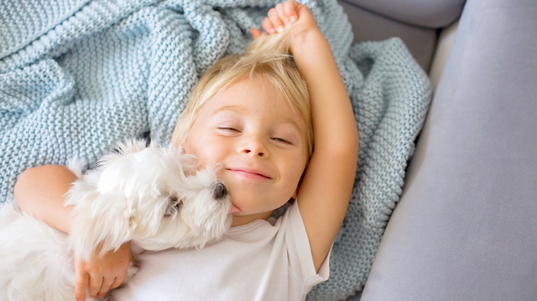 Little kid laying in bed with a dog licking their face.