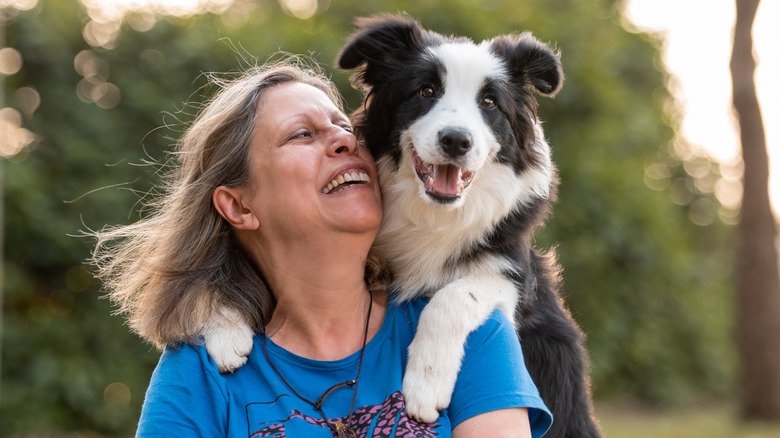 Border collie dog on woman's shoulders posing by the sunset during golden hour green trees in the background.