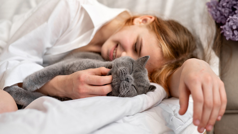 Woman lying down on bed with cat with its eyes closed as she scratches its chin