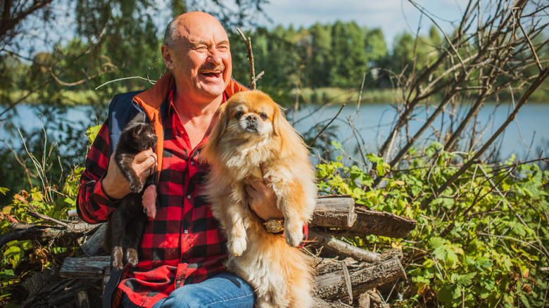 A smiling senior man sitting by the shore with a cat and a shih tzu in his arms.