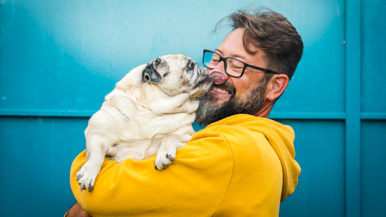 A smiling man in a yellow sweatshirt and glasses hugging a pug.