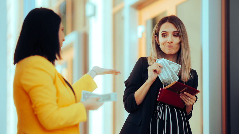 Two business women, one with her hand out, the other with several dollar bills, hesitant to pay
