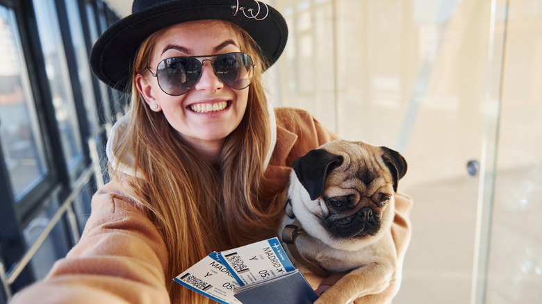 Young white female passenger holding tickets and a pug in an airport hall.