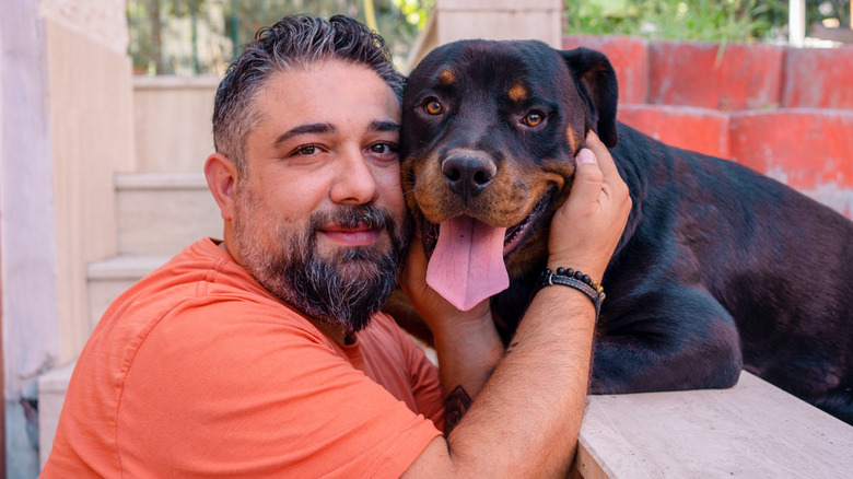 A middle-aged man in an orange short hugging a Rottweiler outdoors.