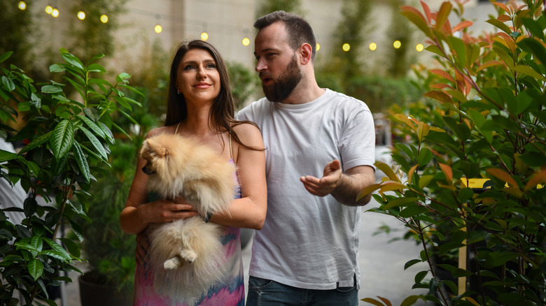 Smiling woman outdoors near plants holding Pomeranian, man standing beside them, talking to her