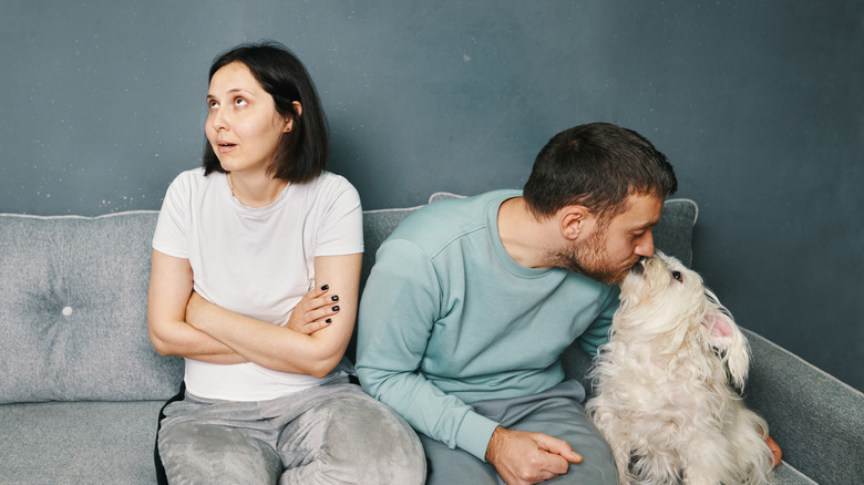 Man on sofa, kissing his dog, woman with arms crossed beside him rolling her eyes