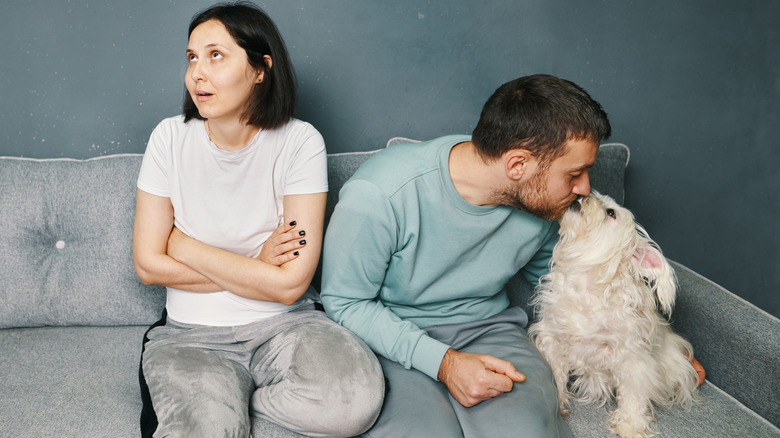 Man on sofa, kissing his dog, woman with arms crossed beside him rolling her eyes